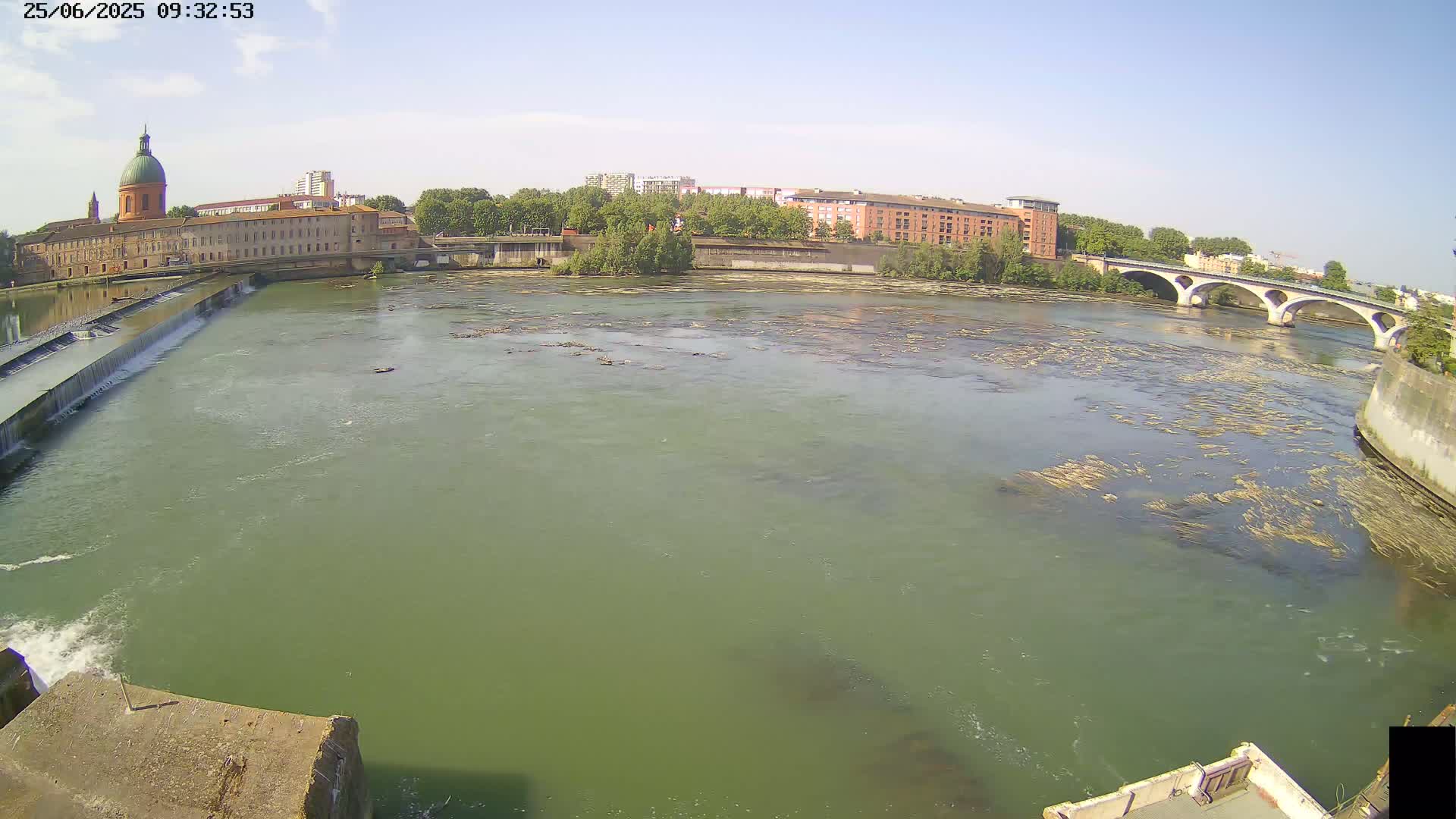 A wide river flows under a stone bridge past a city with buildings and a domed structure, under a clear blue sky.