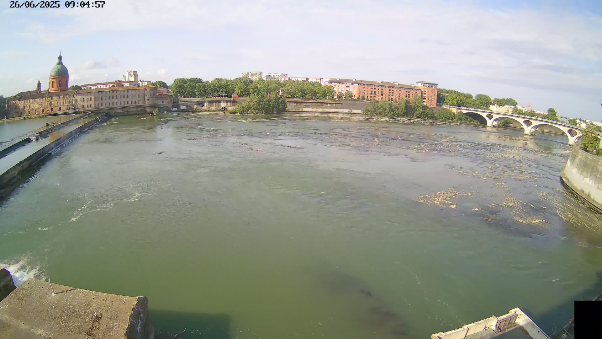 A wide, murky green river flows under a bridge, past a building with a dome and other structures, on a partly sunny day.