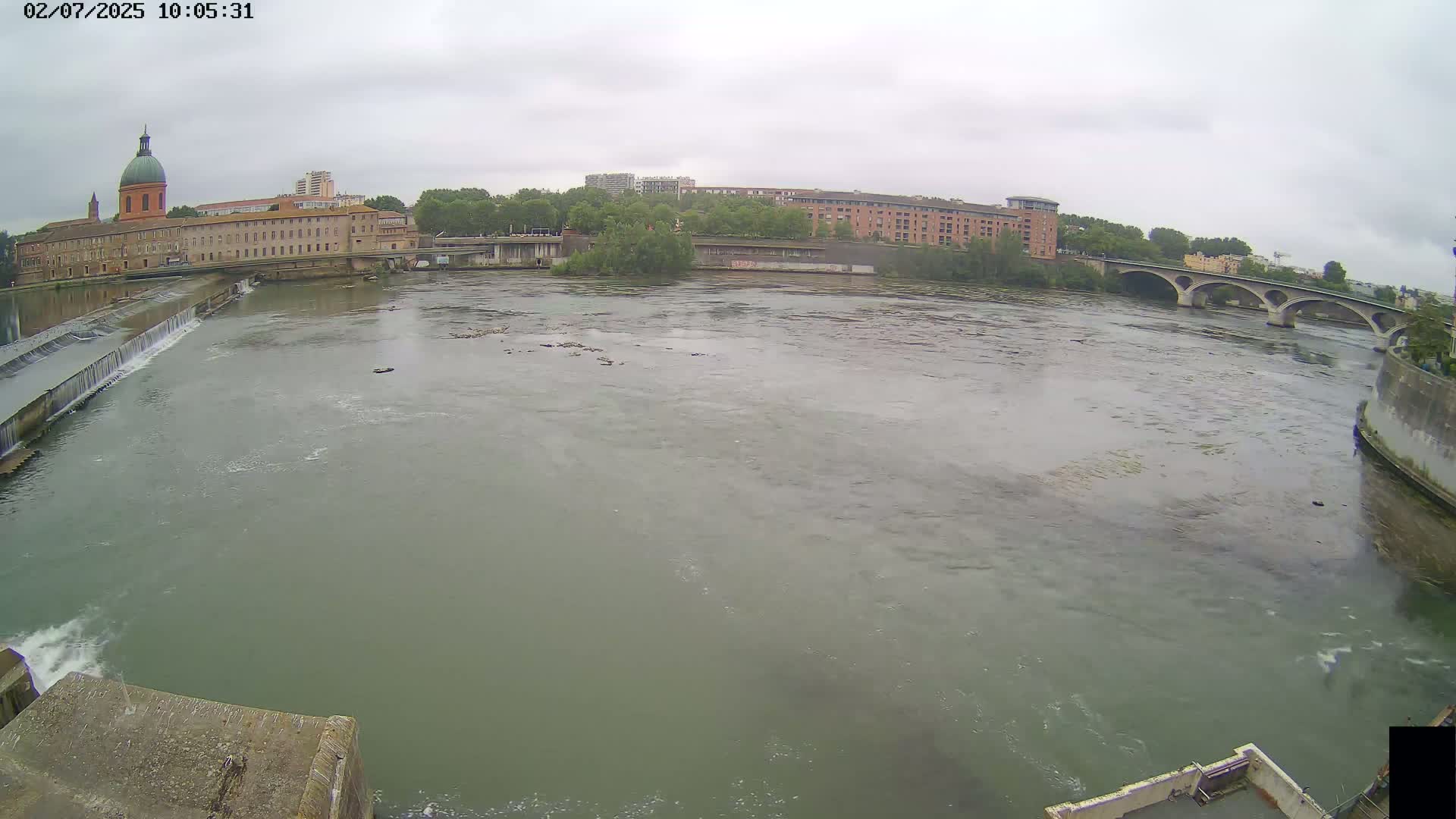 A wide shot of a river on an overcast day shows a building with a dome, other buildings along the shore, a bridge in the distance, and a dam in the foreground.
