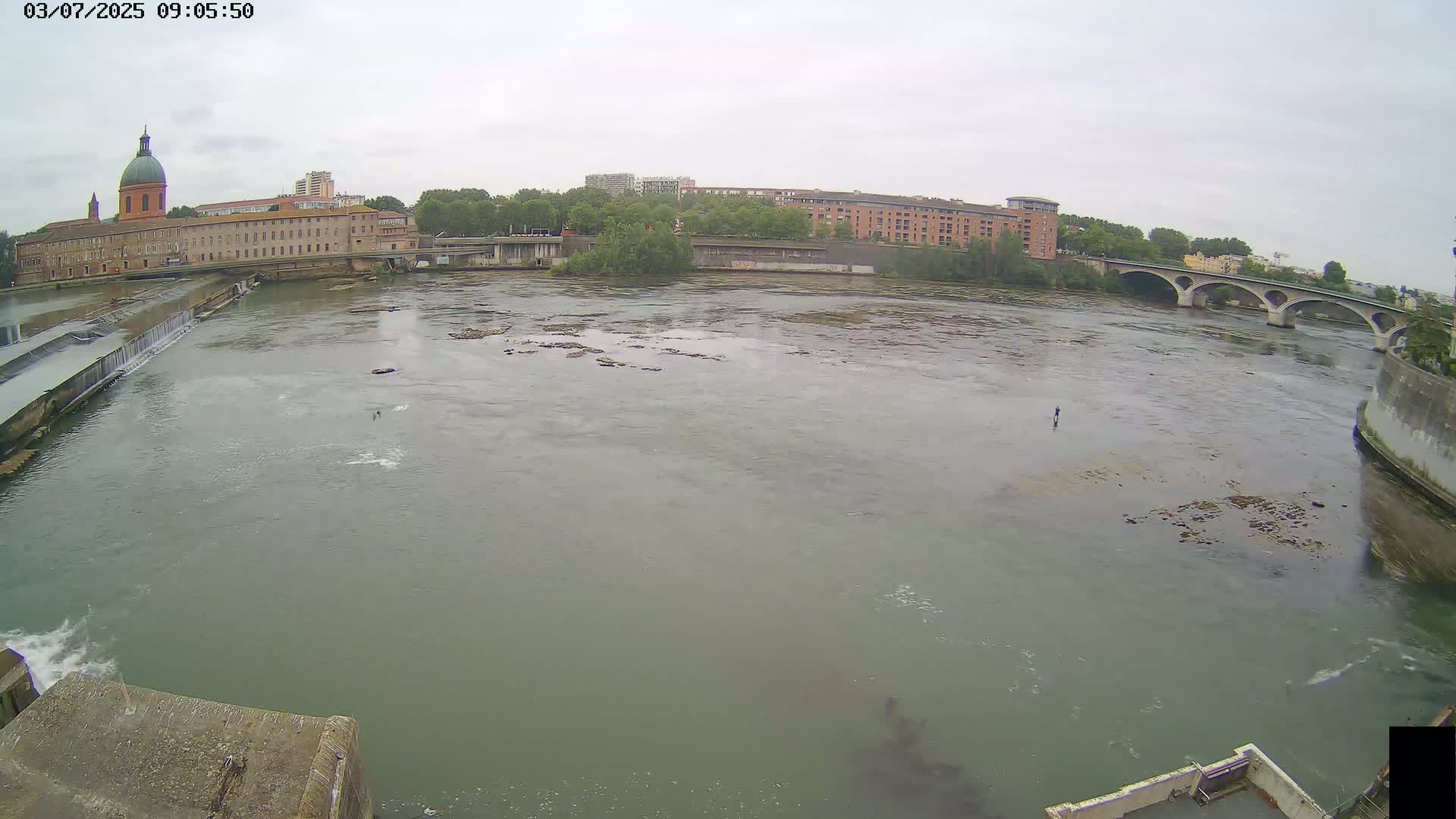 A wide shot of a wide, murky river flowing past a city under an overcast sky shows a dam, a bridge, and several buildings.