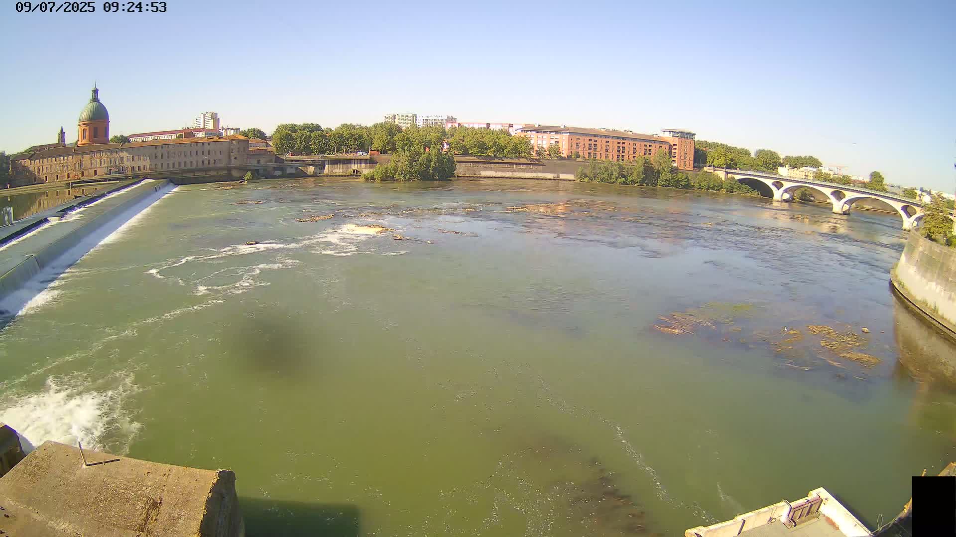 A wide shot of a river flowing past a dam and buildings under a clear blue sky.