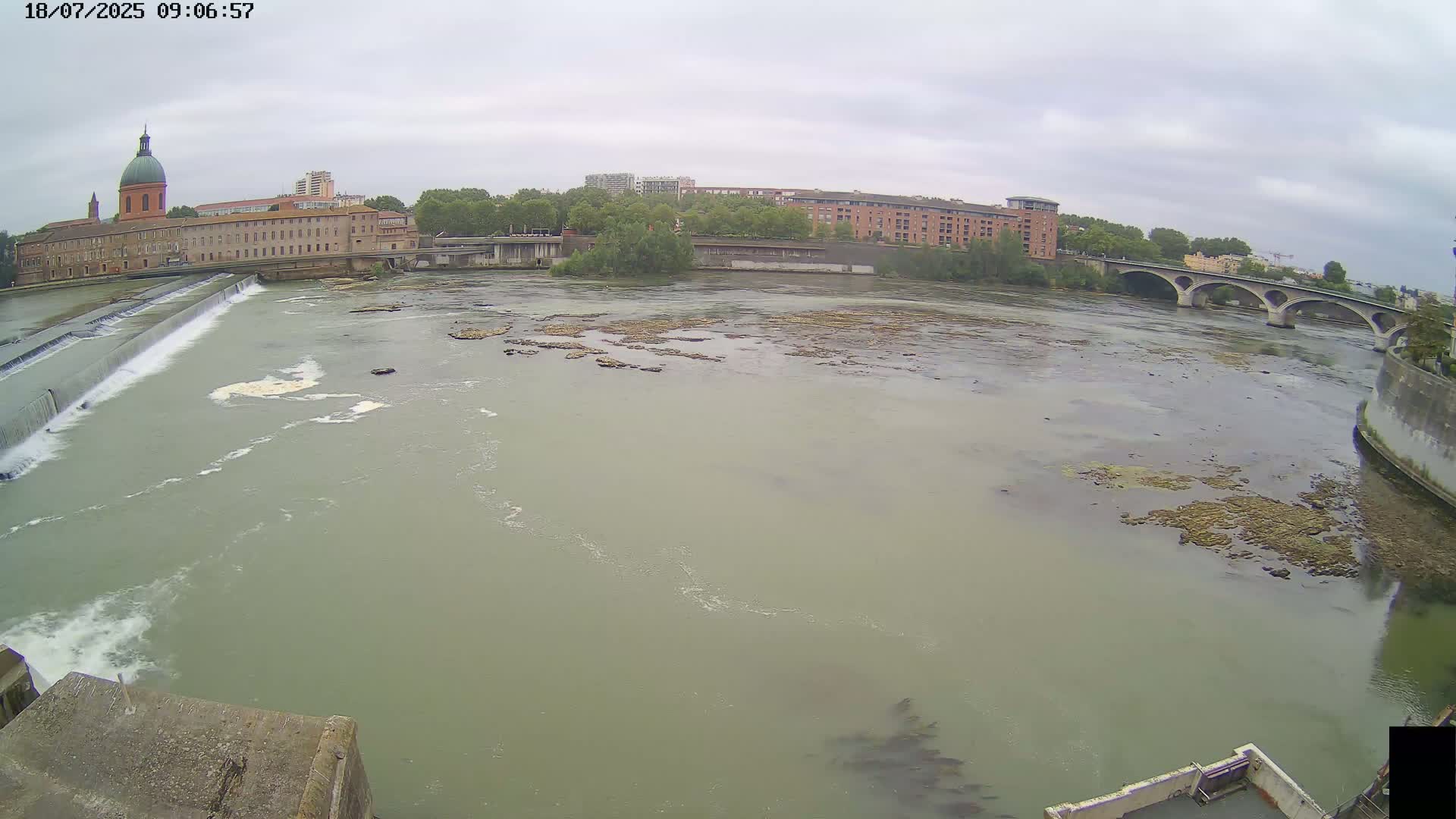 A wide shot of a river flowing past a dam and buildings under an overcast sky.