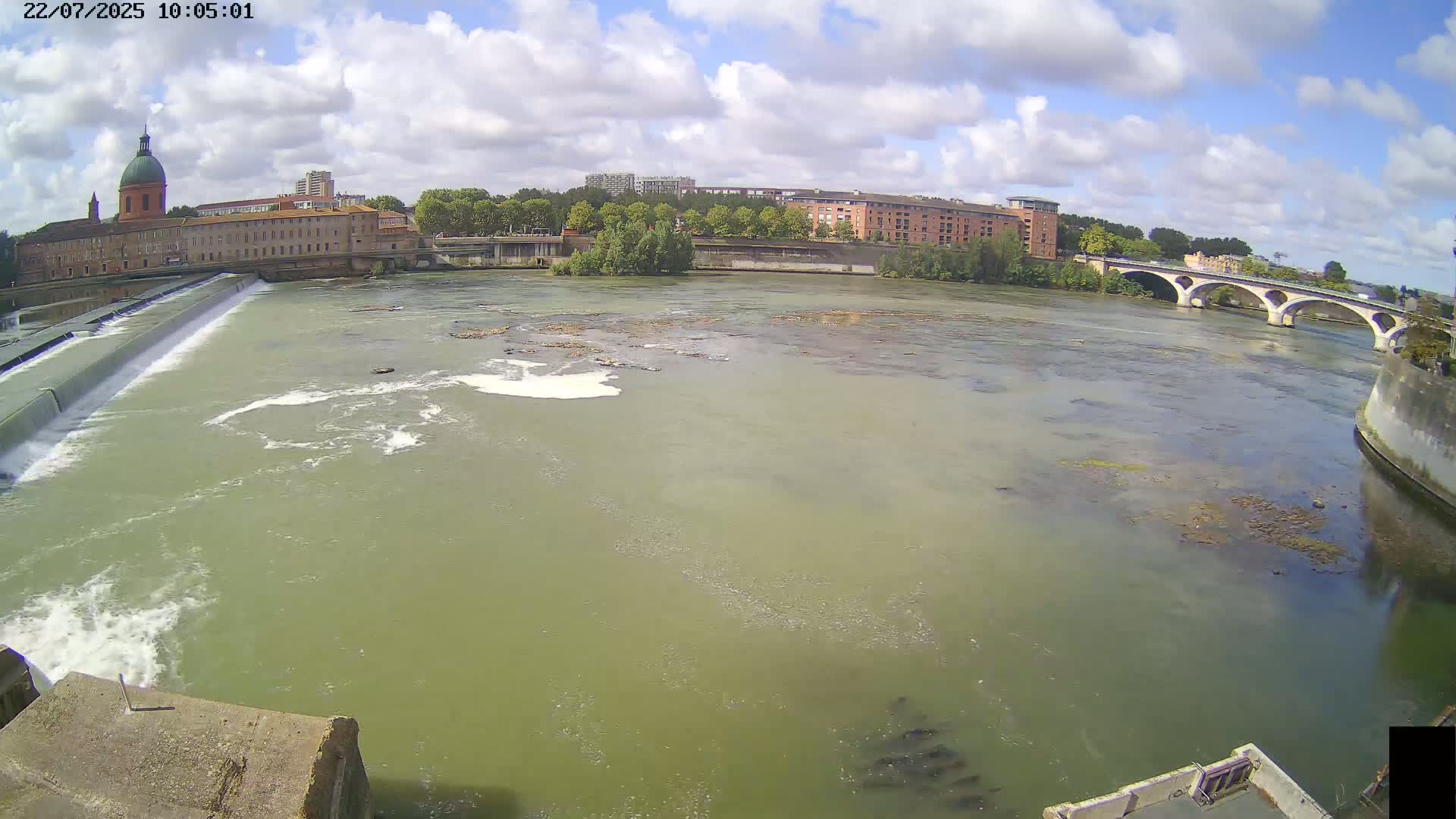 A wide shot of a river with a dam and buildings on the opposite bank under partly cloudy skies.