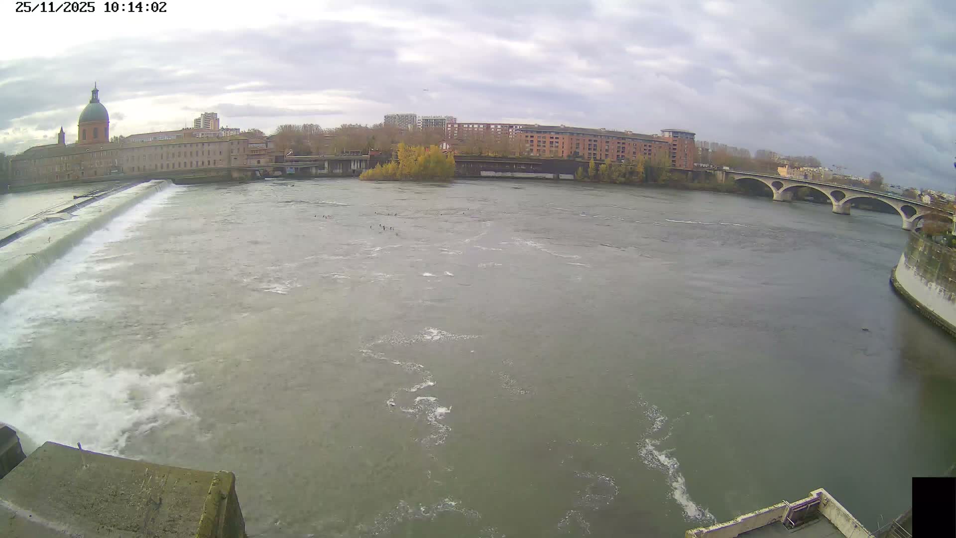 A wide river with a cascading weir flows through an urban landscape featuring a large domed building, multiple bridges, and treed banks, all under a heavily overcast sky.