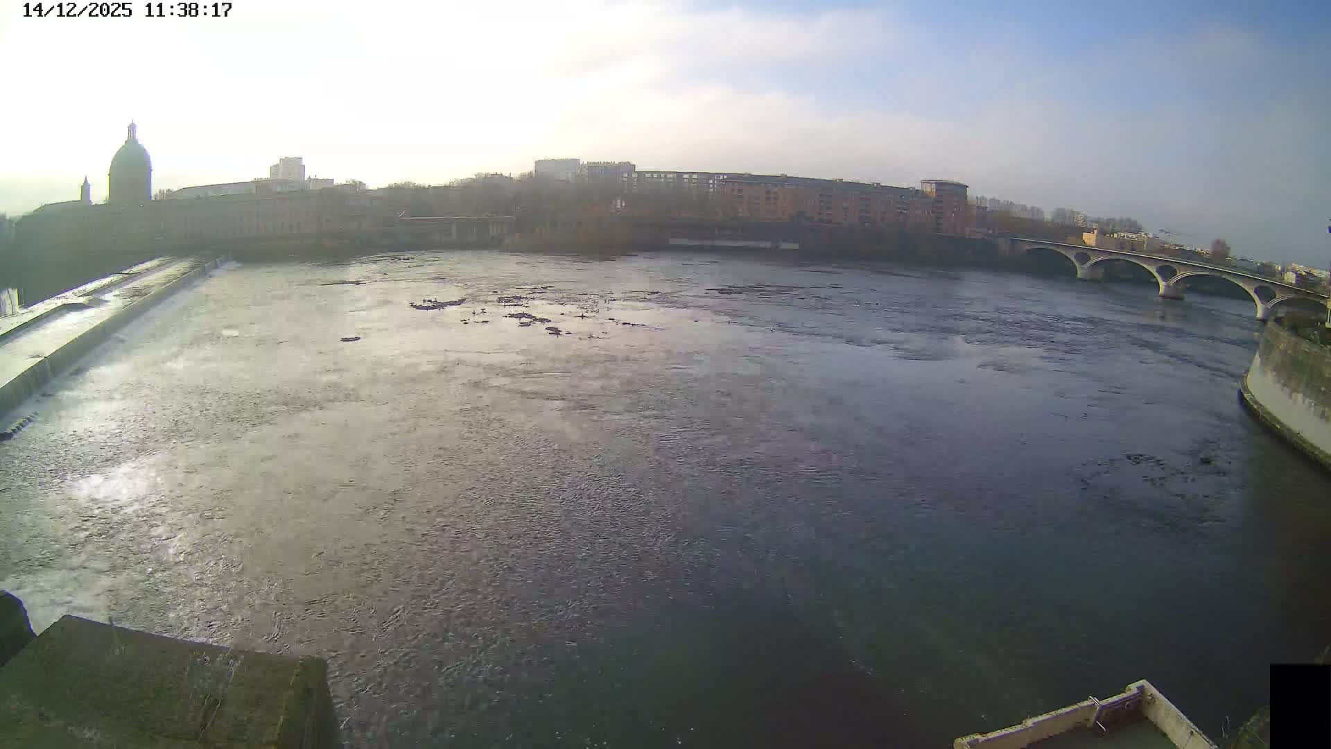A wide river with a cascading weir flows through an urban landscape featuring a large domed building, multiple bridges, and treed banks, all under a heavily overcast sky.