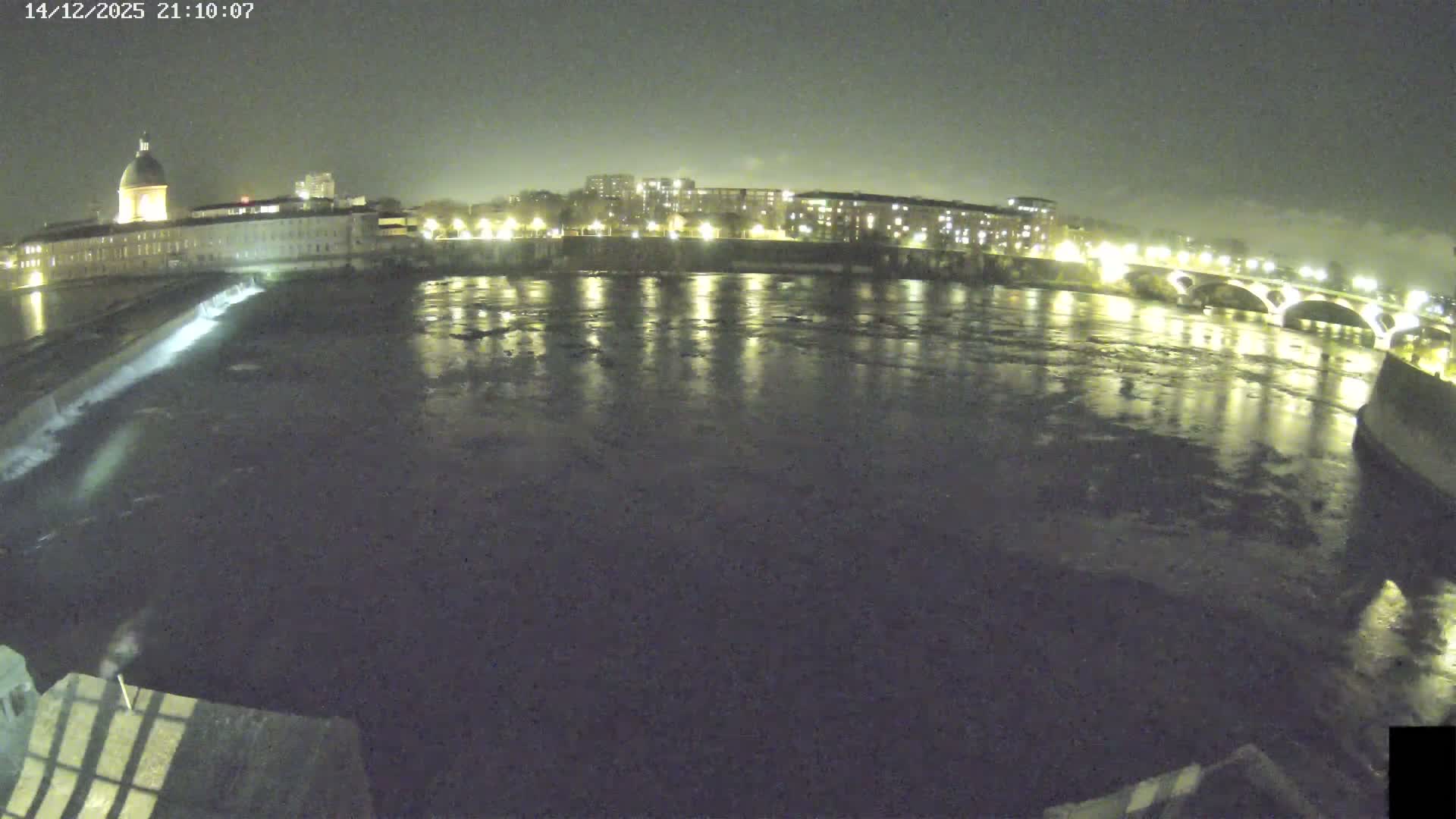 A wide river with a cascading weir flows through an urban landscape featuring a large domed building, multiple bridges, and treed banks, all under a heavily overcast sky.