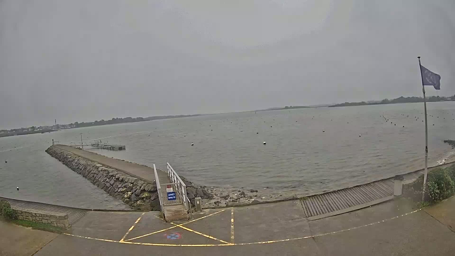 A stone jetty extends into a calm, grey body of water under an overcast sky, with a small boat ramp and a flagpole visible on the shore.