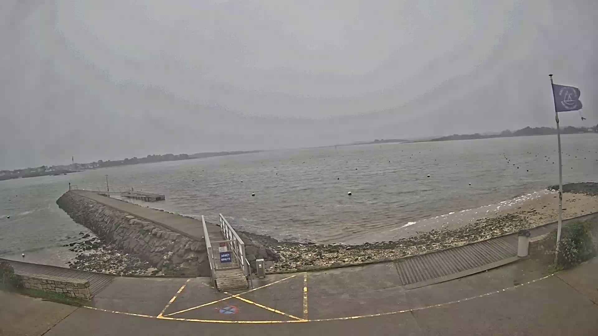 A stone jetty extends into a calm, grey body of water under an overcast sky, with a sandy shore and a ramp leading to a paved area.