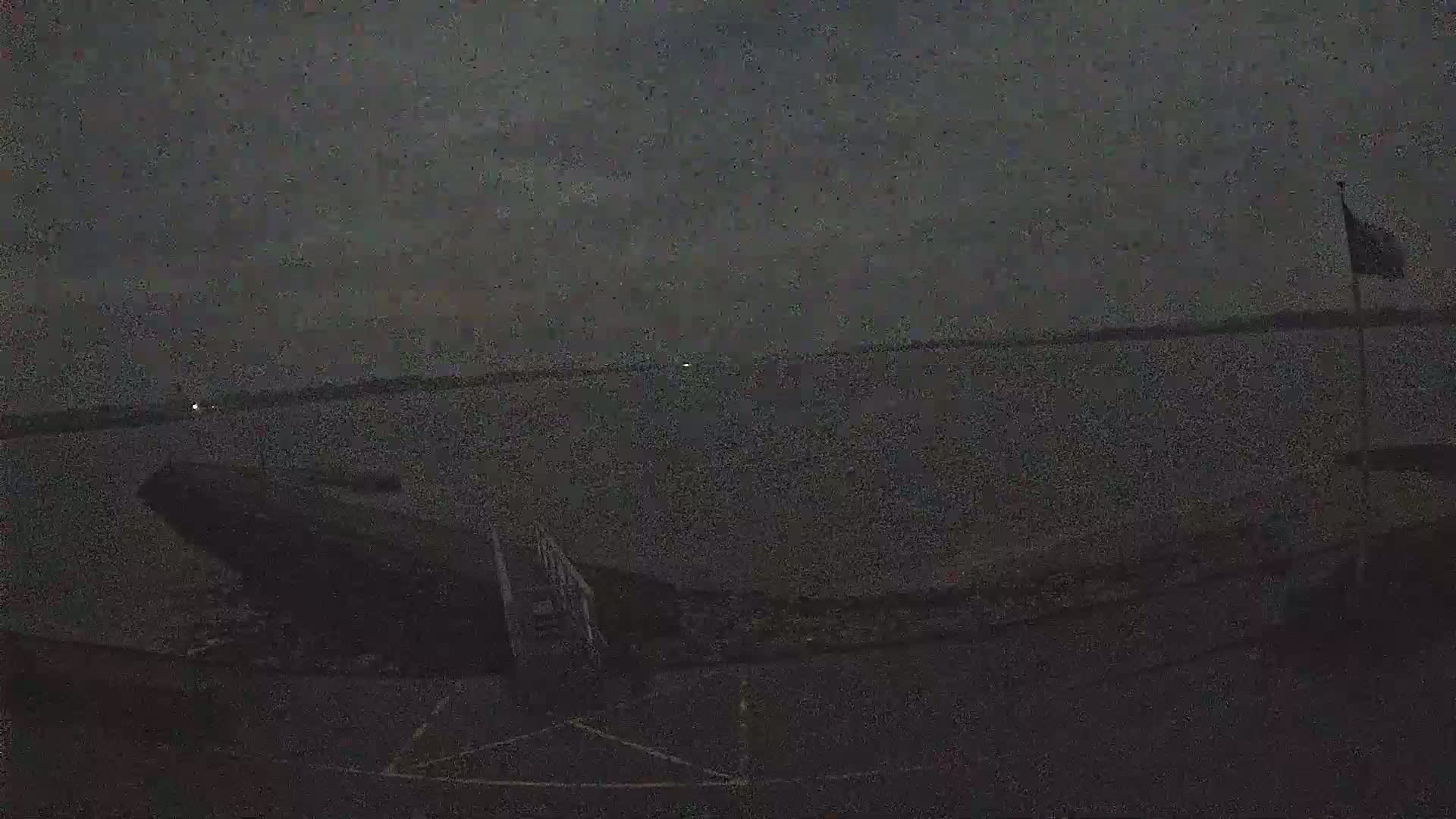 A dark, possibly nighttime, scene shows a body of water under a mostly cloudy sky, with a partially visible structure and a flagpole in the foreground.