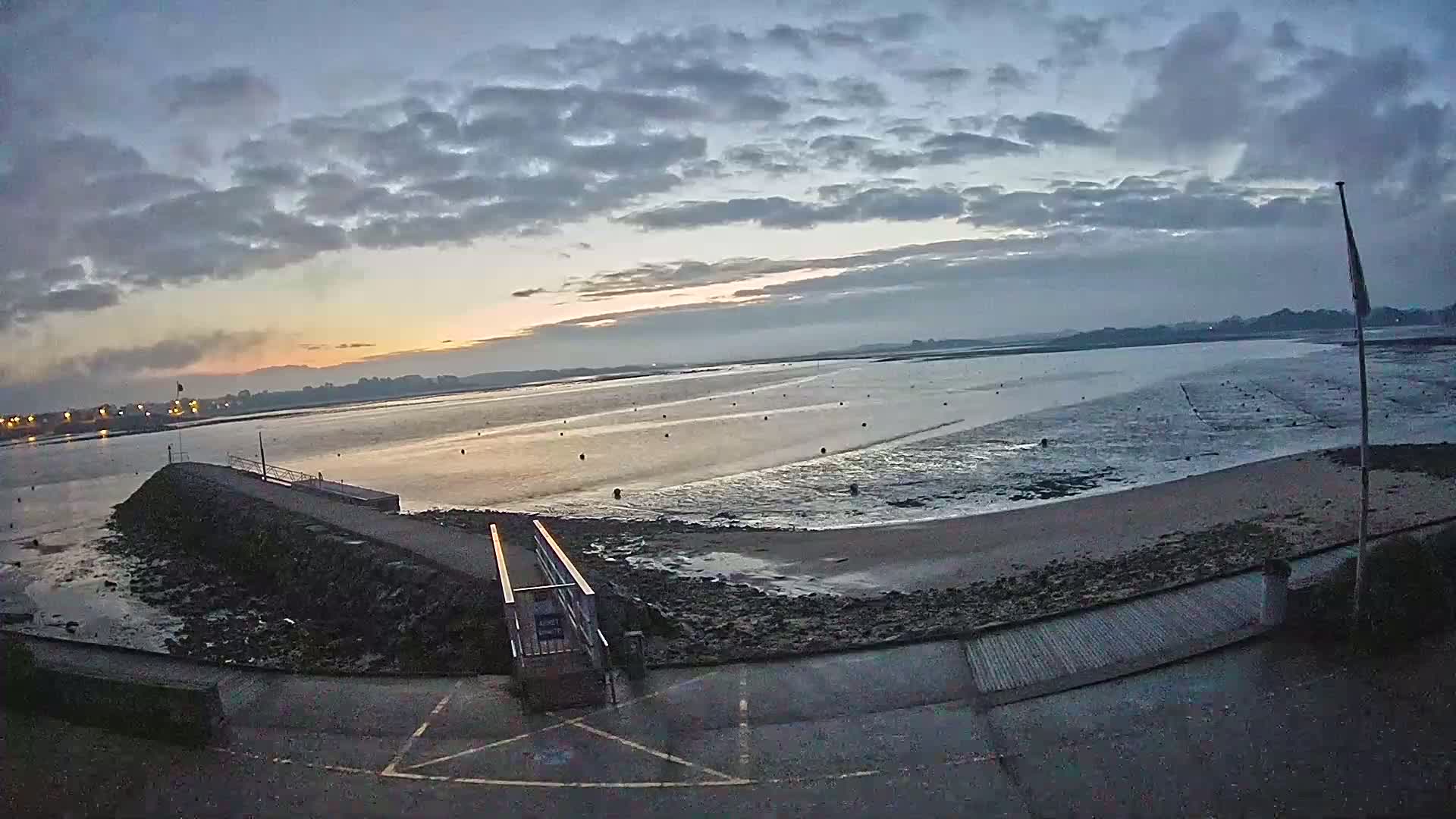 A calm, overcast sunrise over a bay with a low-tide sandy beach and a stone pier leading to a ramp.