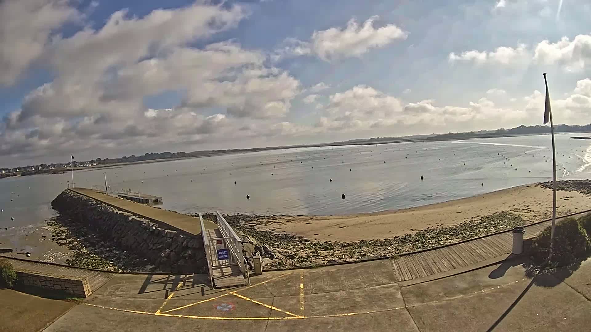A stone jetty extends into a calm bay under a partly cloudy sky, with a sandy beach and numerous dark buoys in the water.