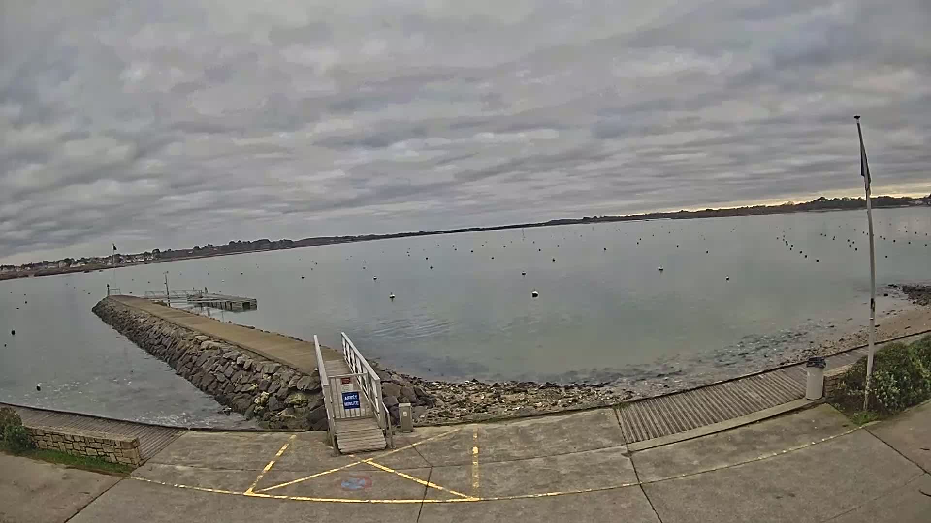 A stone jetty extends into calm, grey water dotted with buoys under a cloudy sky.