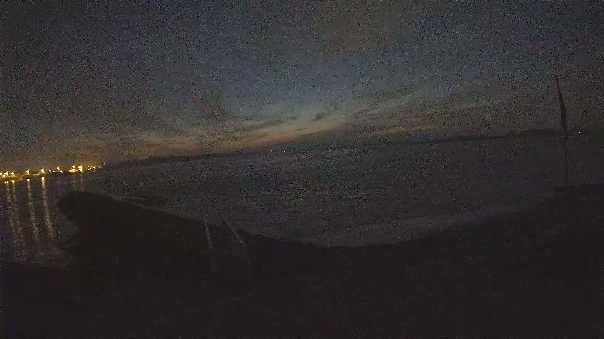 A dark, nighttime scene shows a body of water with lights in the distance under a partly cloudy sky.