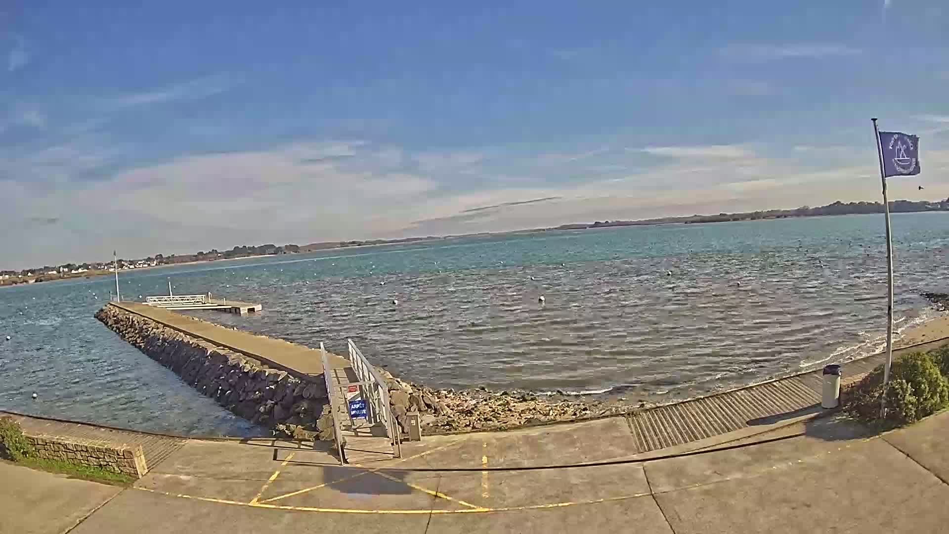 A stone jetty extends into a calm, teal-colored body of water under a mostly sunny sky with a few clouds.