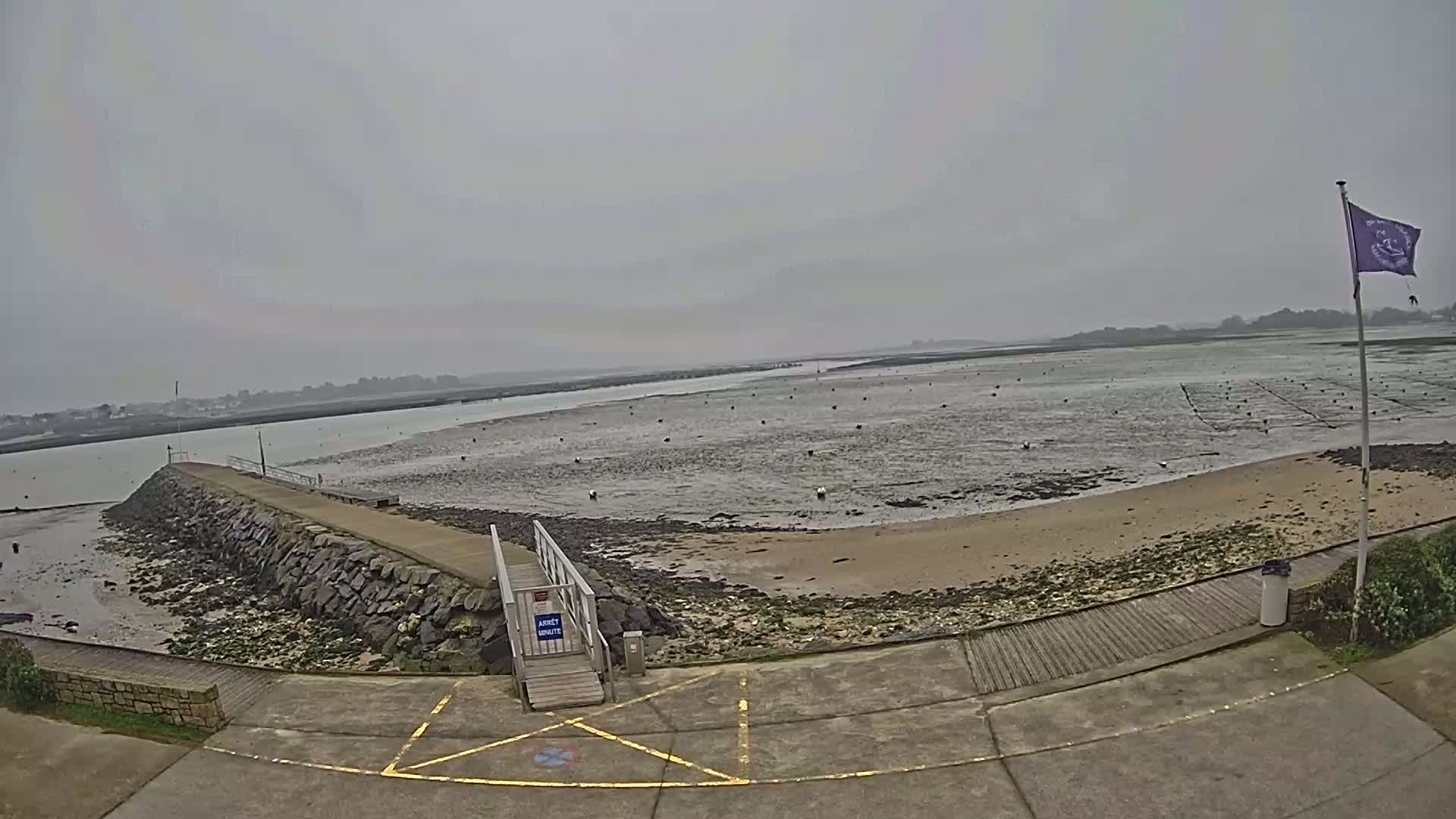 A stone jetty extends into a flat, muddy expanse of water under an overcast sky, with a sandy beach and oyster beds visible in the distance.