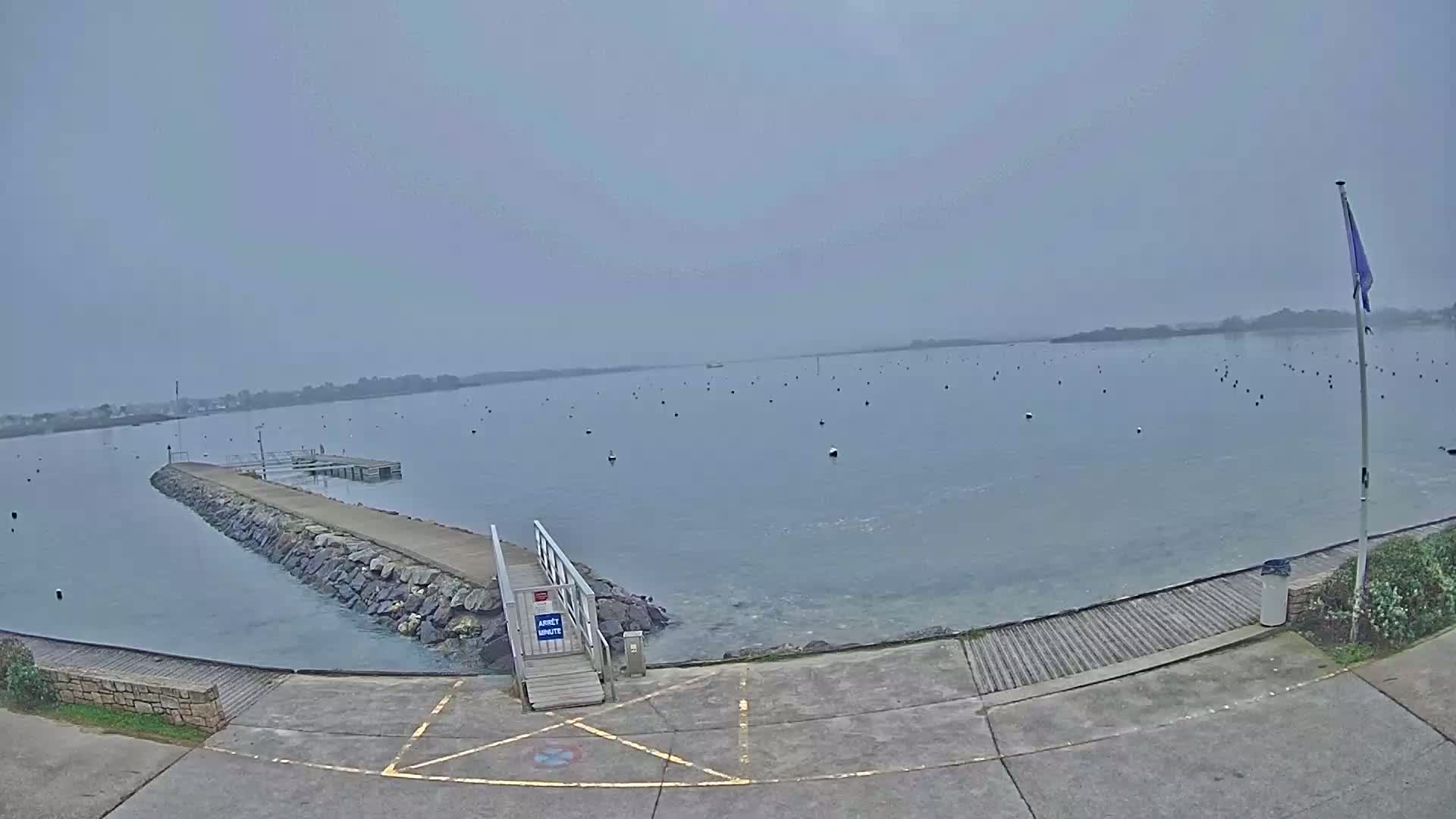 A calm, gray day shows a stone jetty extending into a body of water with numerous dark buoys scattered across its surface, a small wooden dock is visible at the end of the jetty.