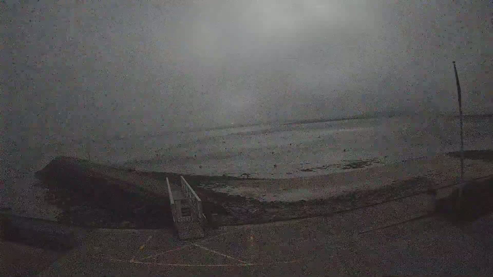A dark, overcast day reveals a beach with a small building near a walkway leading to the water, where a few people are visible.