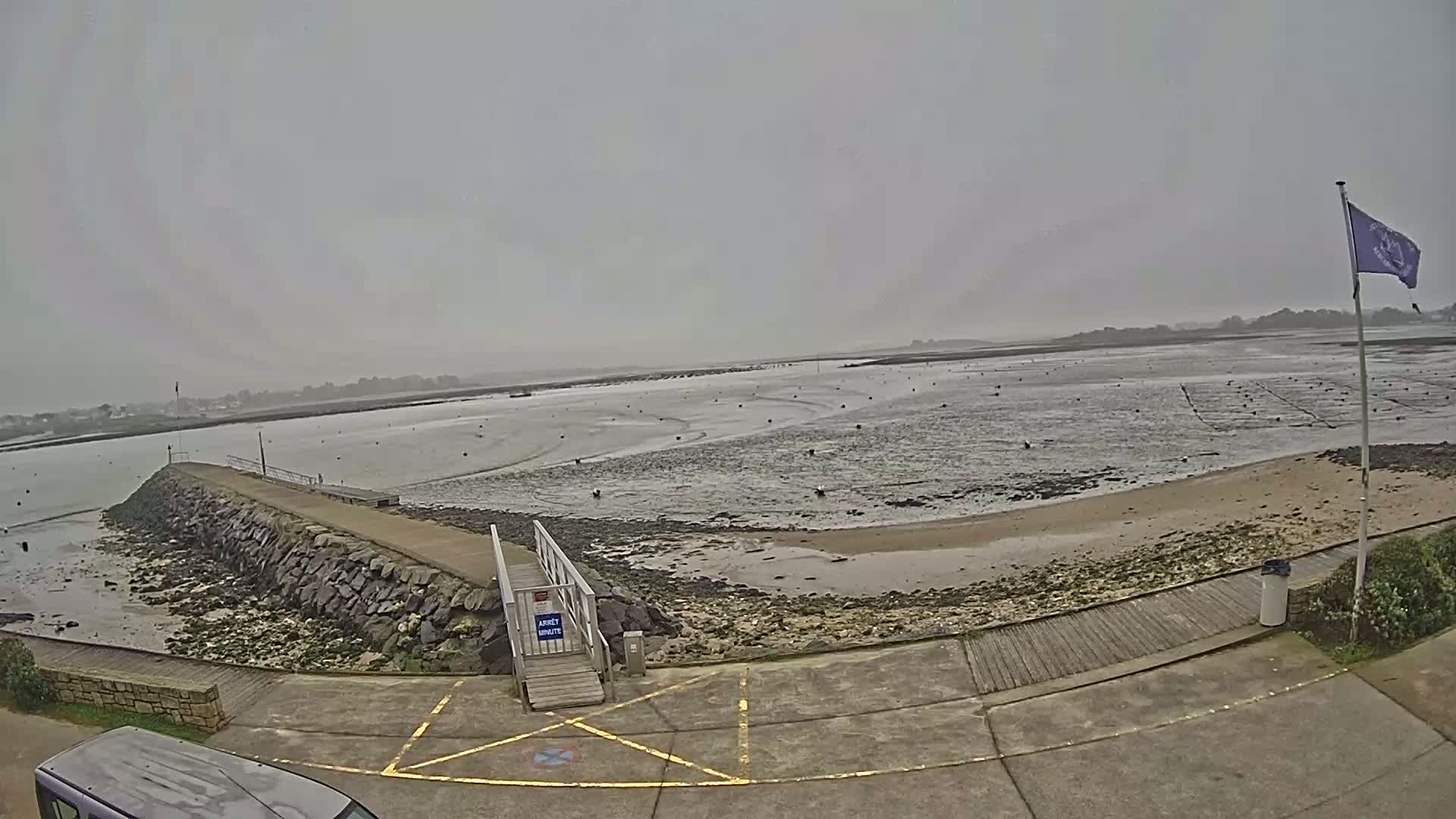 A low-tide coastal scene on an overcast day shows a stone jetty leading to a sandy beach with a wooden walkway and a flagpole.