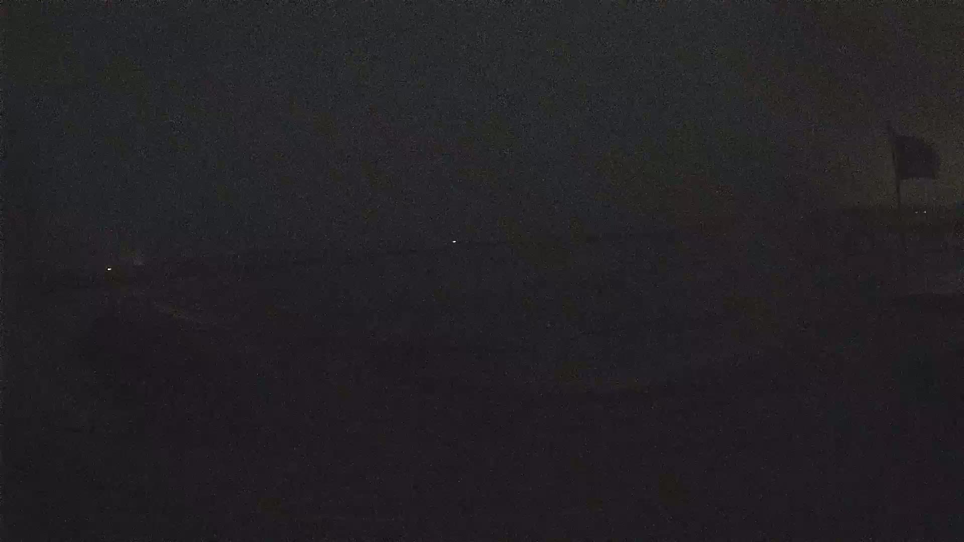 A dark, nighttime scene shows a few distant lights on a flat horizon under a mostly clear, dark sky.