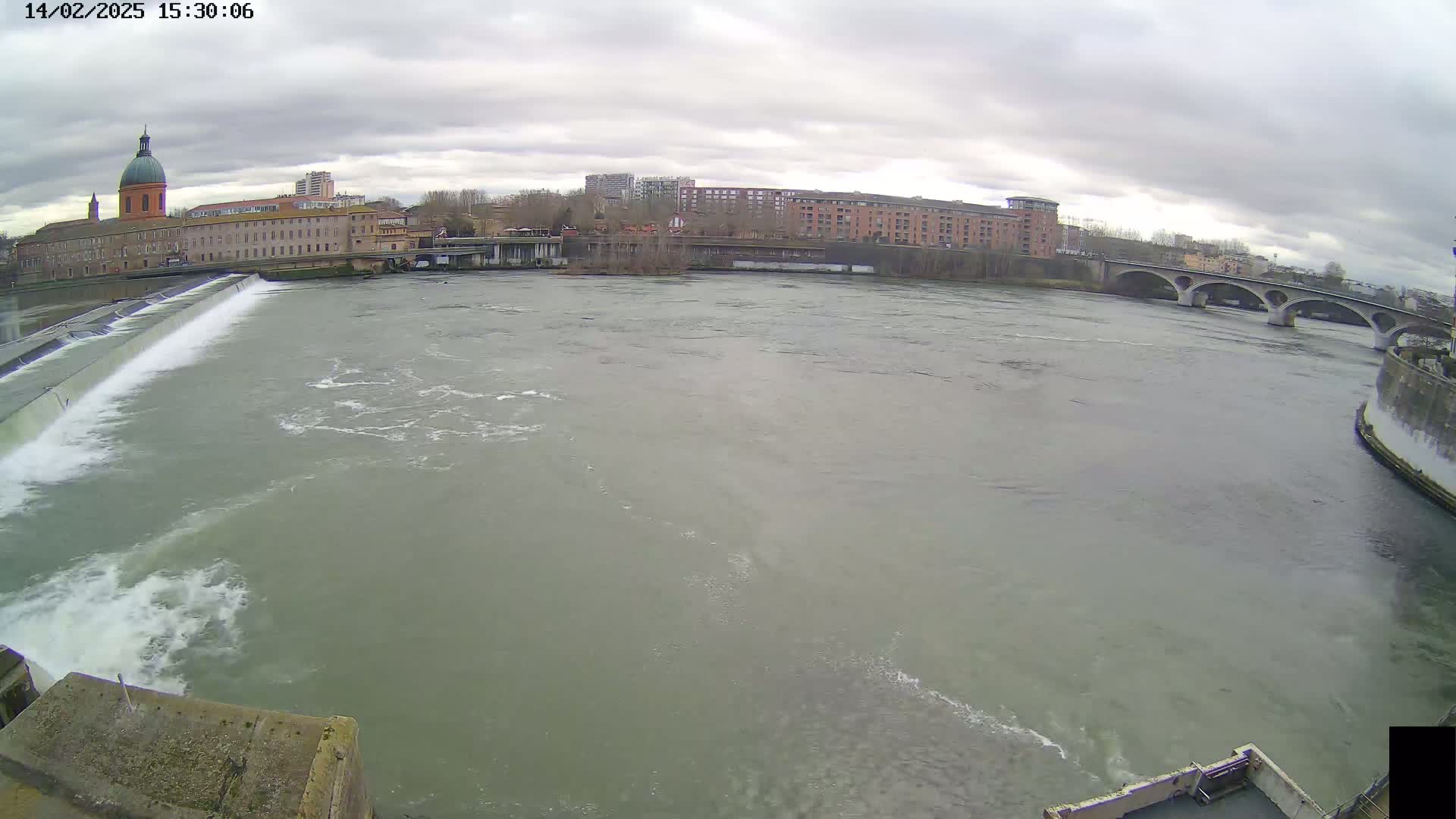 A wide shot shows a river flowing under an overcast sky past a dam and a bridge, with buildings visible in the background.