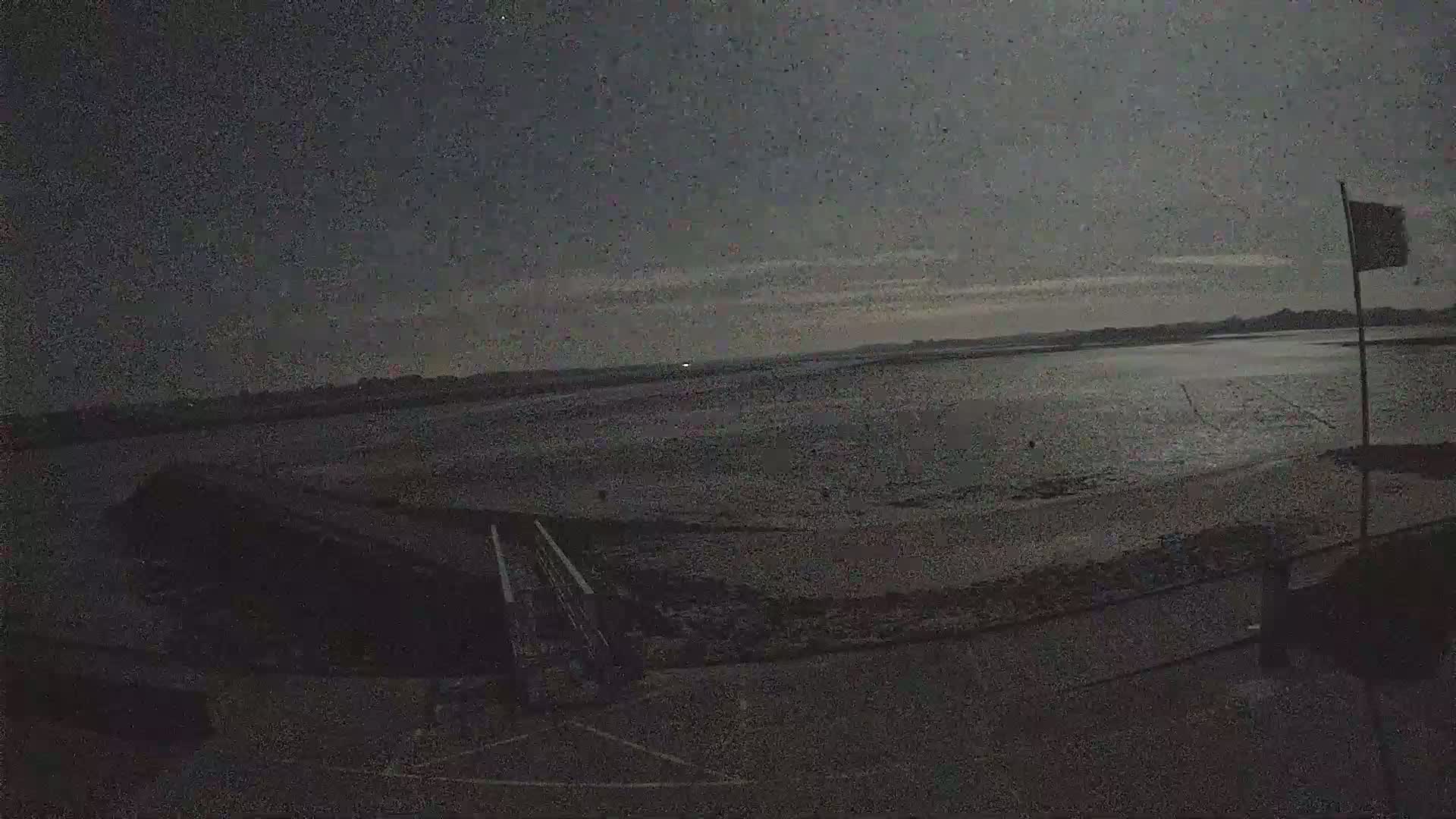 A dark, overcast sky overlooks a wide expanse of flat, dark-colored land meeting a calm body of water under low light conditions.