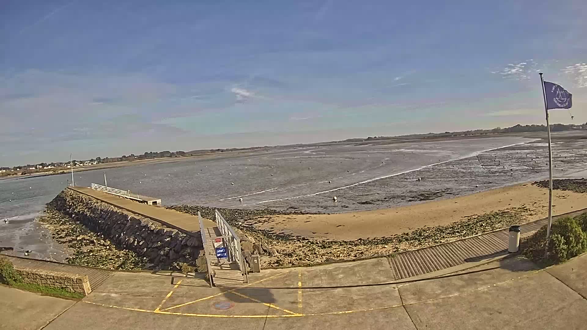 A stone pier extends into a calm, shallow bay on a sunny day, leading to a sandy beach.