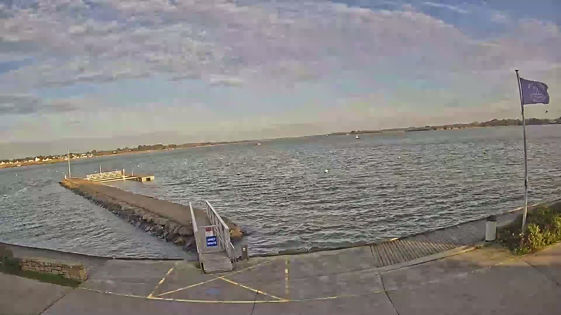 A small stone pier extends into a calm body of water under a partly cloudy sky, with a few boats visible in the distance.