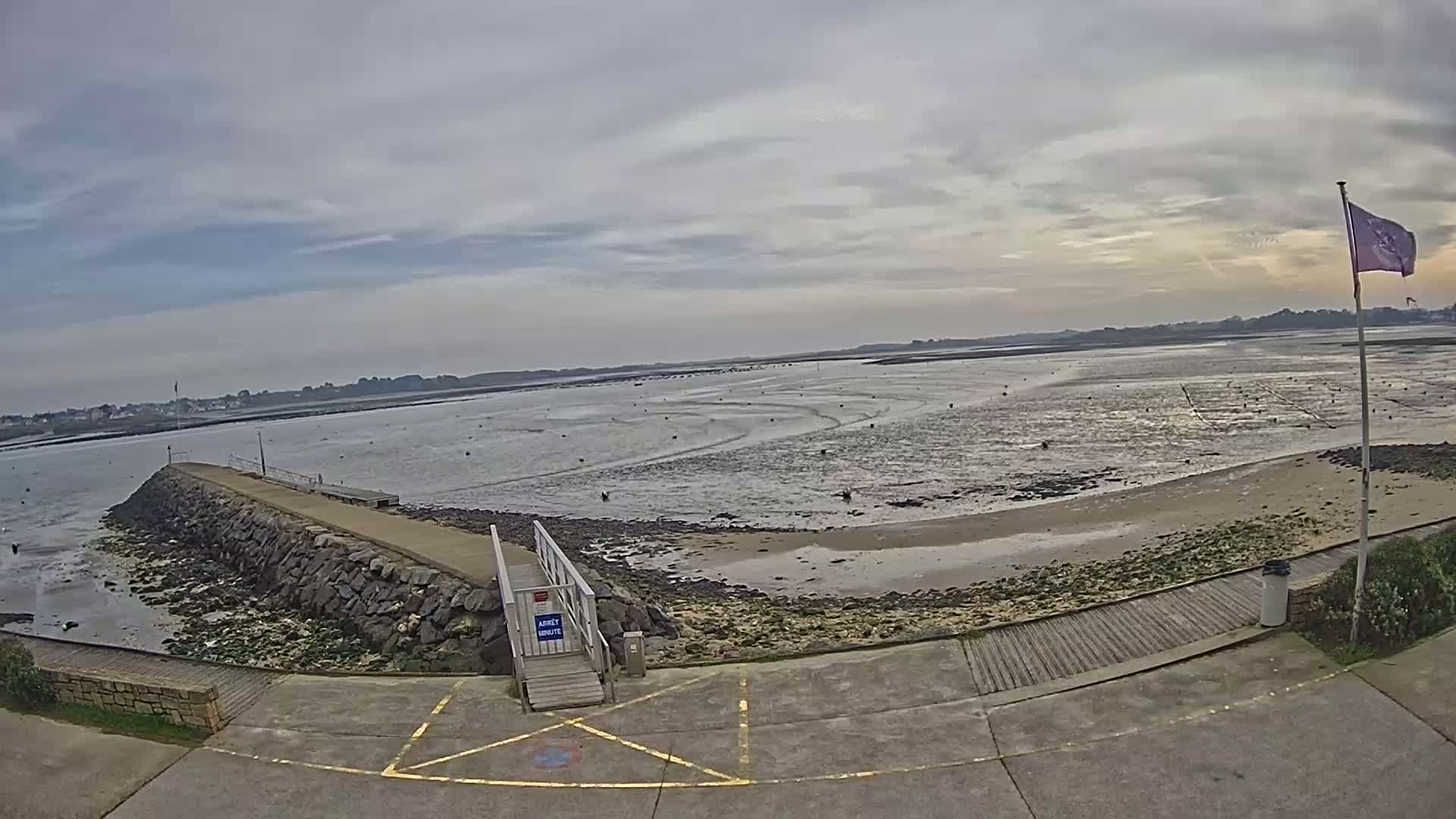 A stone jetty extends into a calm, shallow bay under a cloudy sky, with a sandy beach and low tide visible.