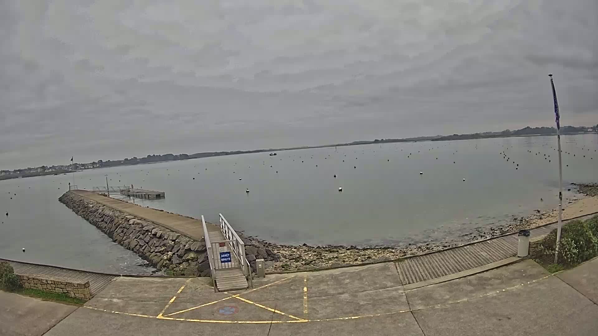 A stone jetty extends into calm, gray water dotted with numerous buoys under an overcast sky.