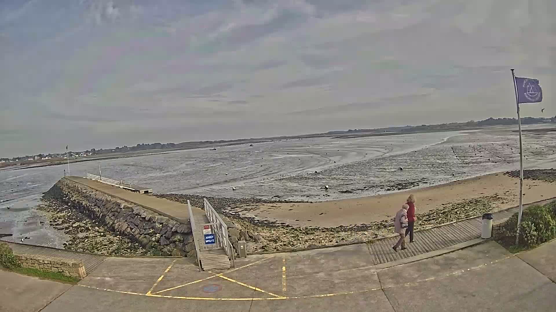 A stone jetty extends into a calm, shallow bay under a mostly cloudy sky, with two people walking along a boardwalk on the shore.
