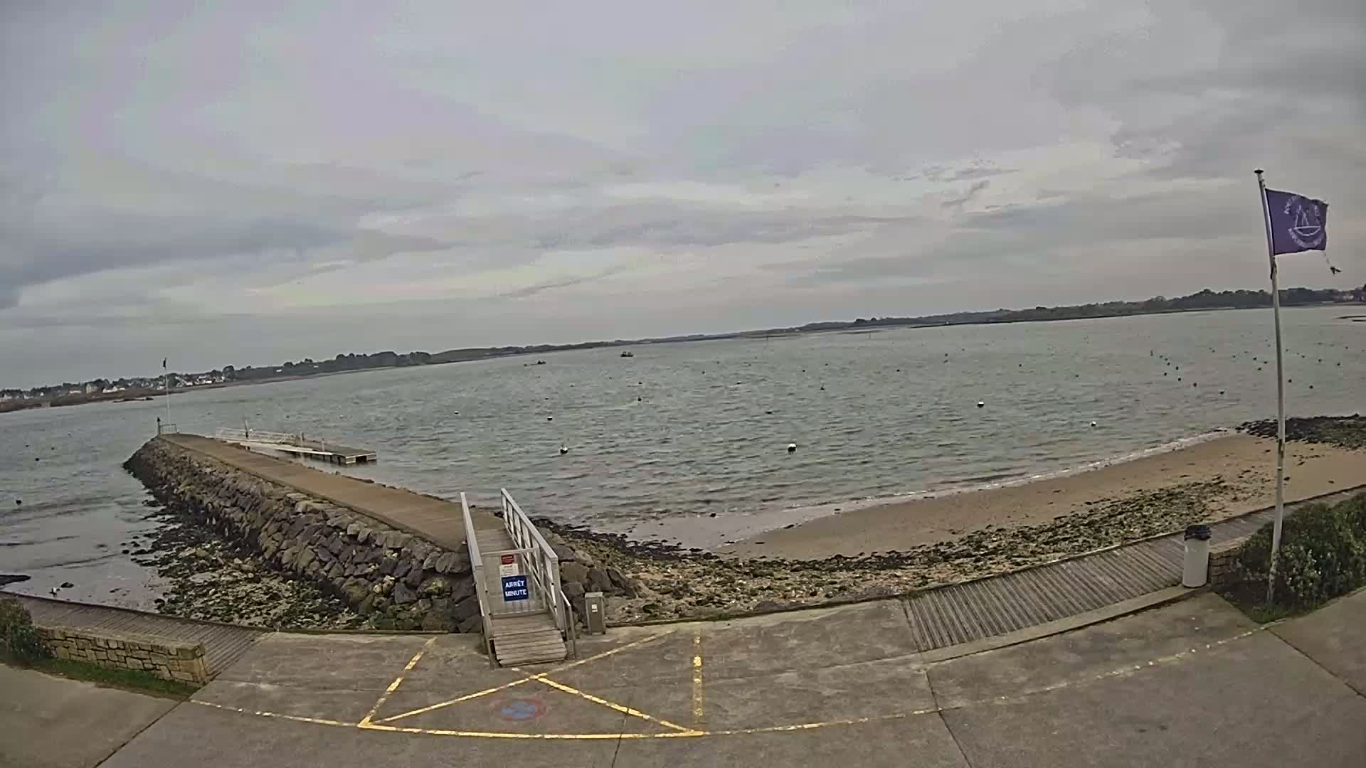 A stone jetty extends into a calm body of water under an overcast sky, leading to a small sandy beach.