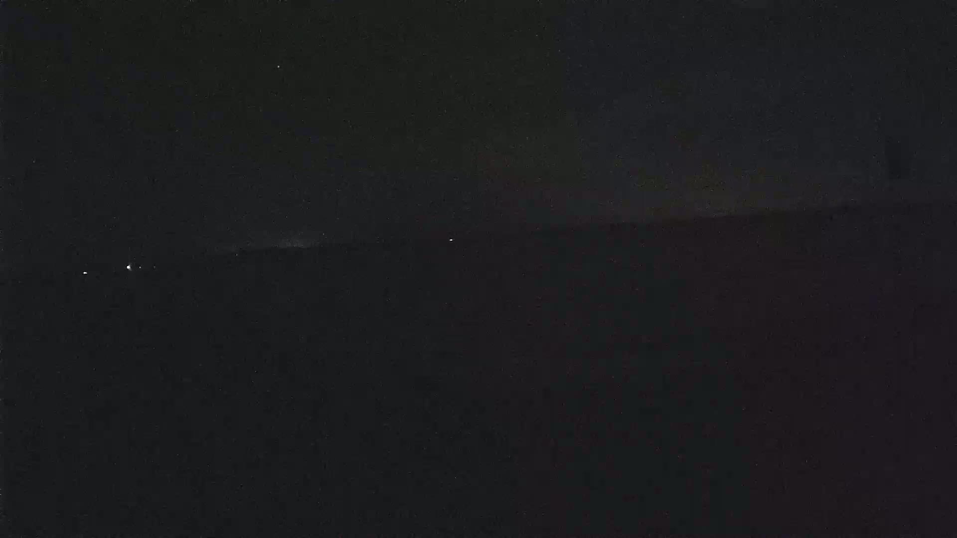 A dark, nighttime landscape with a few distant lights visible on the horizon under a mostly clear sky.