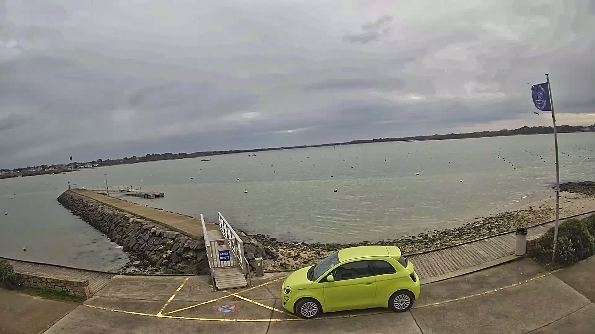 A lime-green car is parked near a stone jetty extending into a calm body of water under an overcast sky.