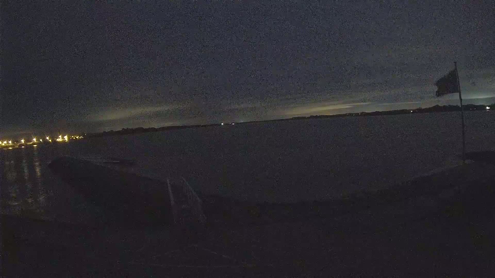 A dark, nighttime view of a body of water with a flagpole and distant lights visible across the water under a mostly clear sky.