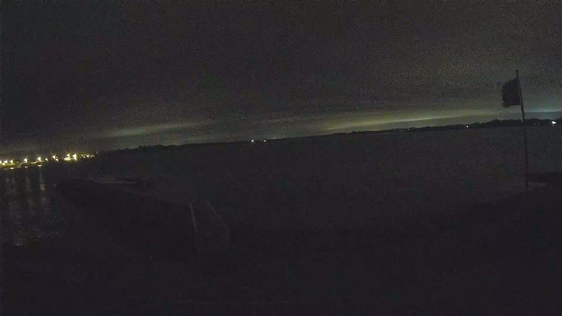 A dark nighttime scene shows a body of water with a distant shoreline under a mostly clear, dark sky; a flagpole with a flag is visible on the right.
