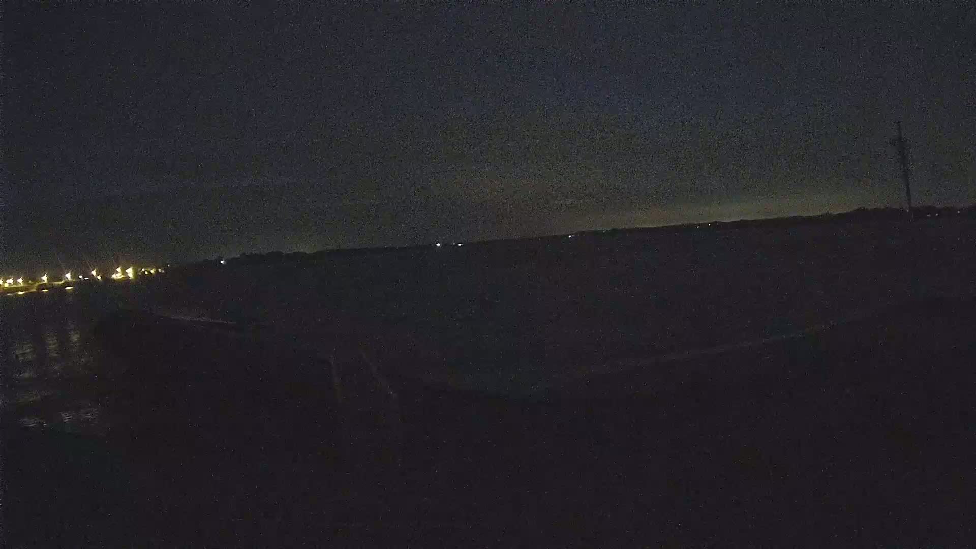 A dark, nighttime scene shows a body of water with a distant shoreline faintly illuminated by lights under a mostly clear sky.