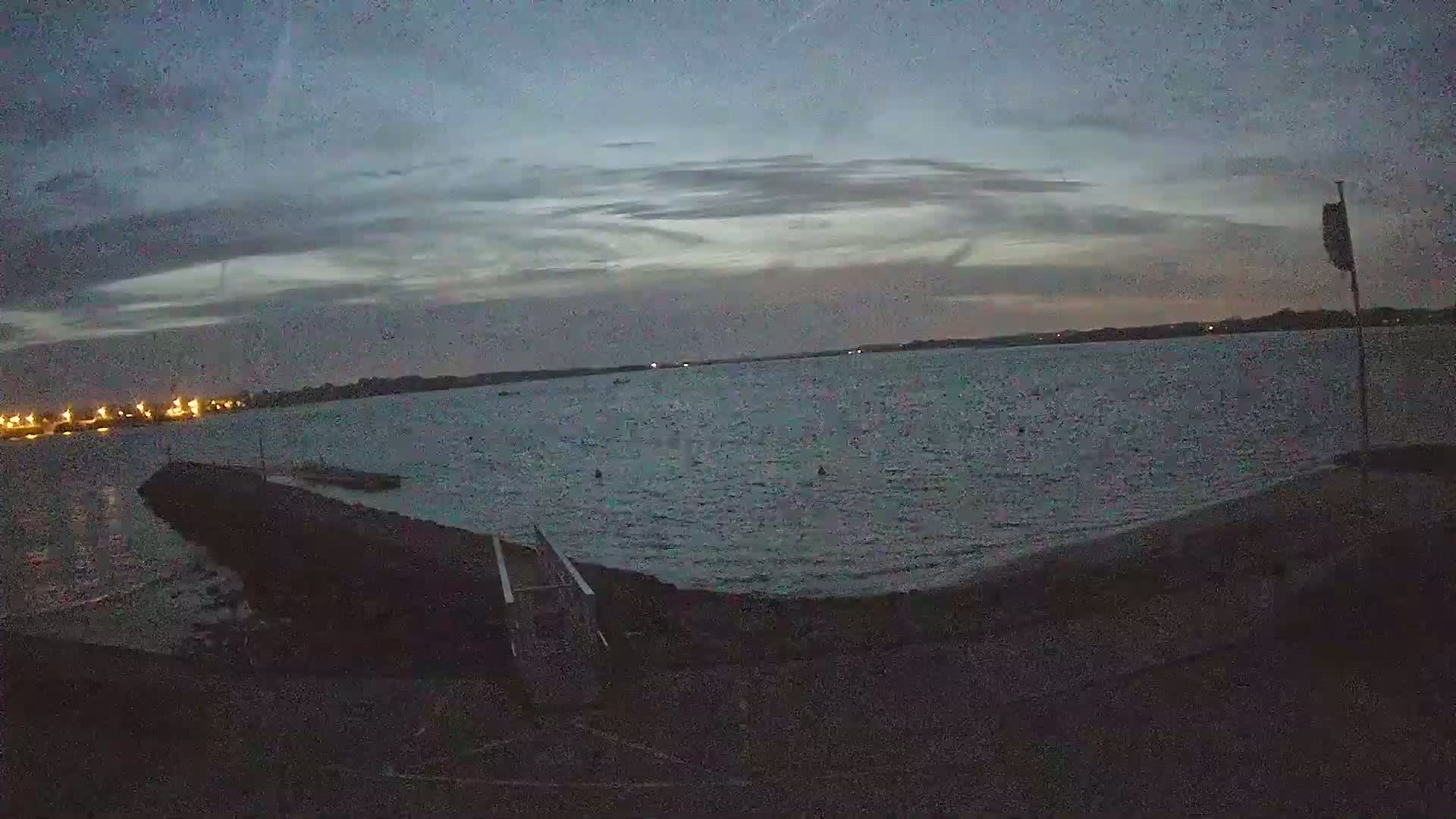 A dark, calm body of water is seen at dusk under a mostly cloudy sky, with a small pier or jetty extending into the water from a darkened shoreline.