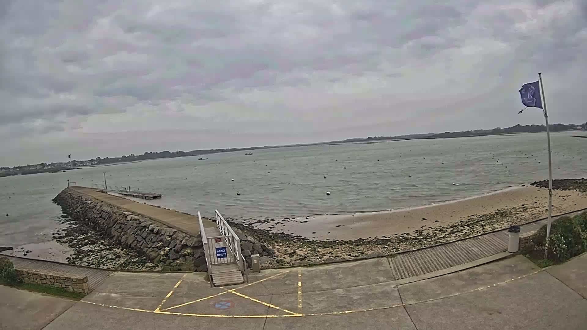 A stone jetty extends into a calm, grey-watered bay under an overcast sky, leading to a small sandy beach.