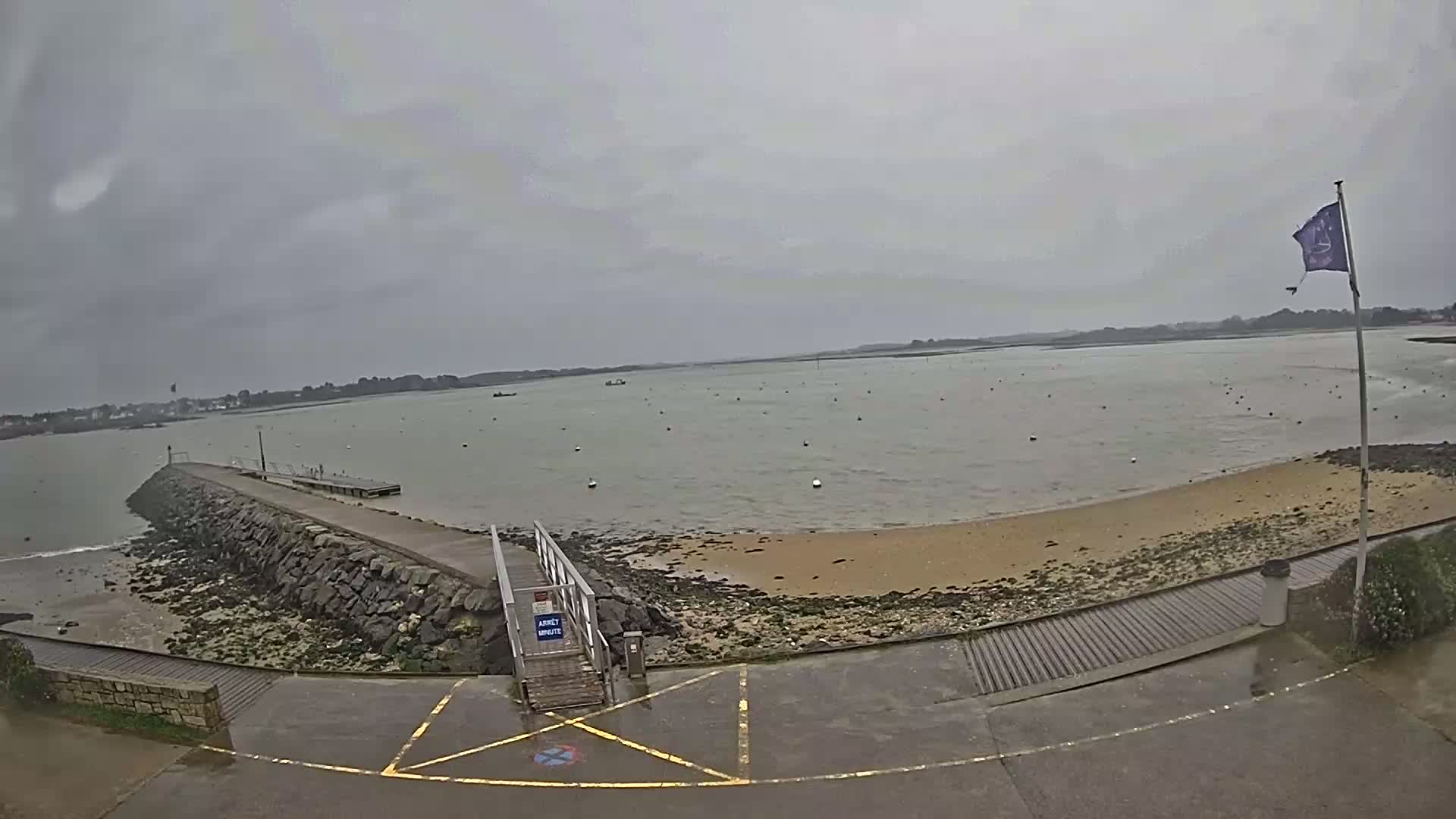 A stone pier extends into a calm, gray body of water under an overcast sky, leading to a small sandy beach.