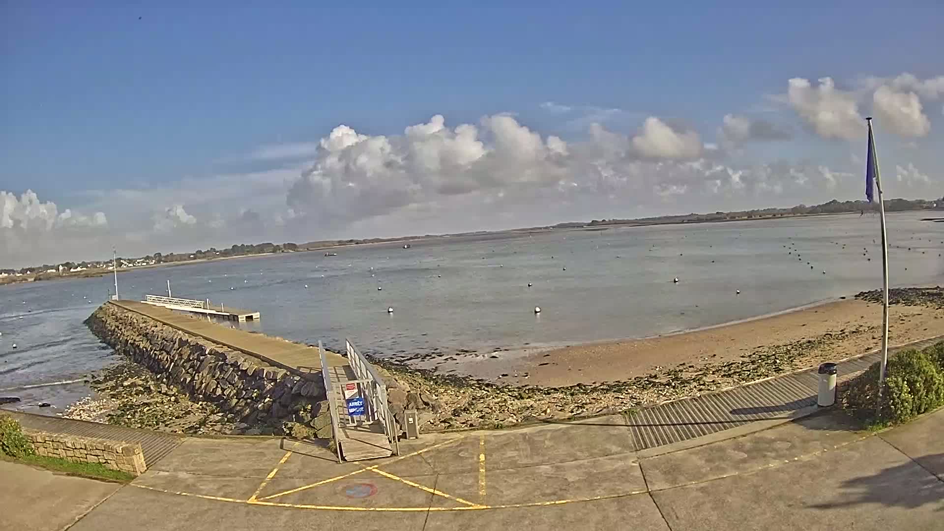 A stone pier extends into a calm body of water under a partly cloudy sky, with a sandy beach visible to the right.
