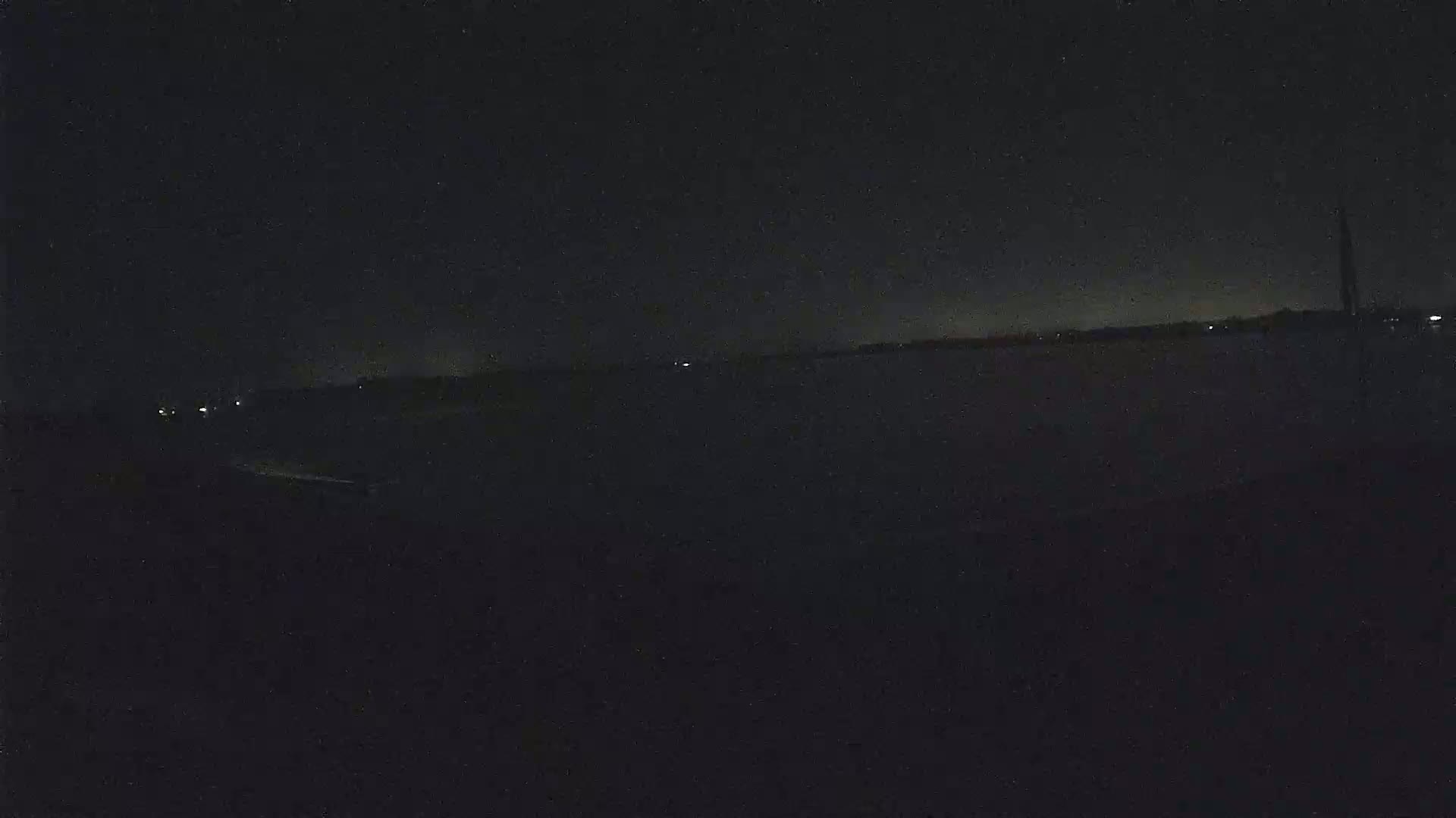A dark, nighttime view of a flat landscape with a few distant lights under a clear, starless sky.