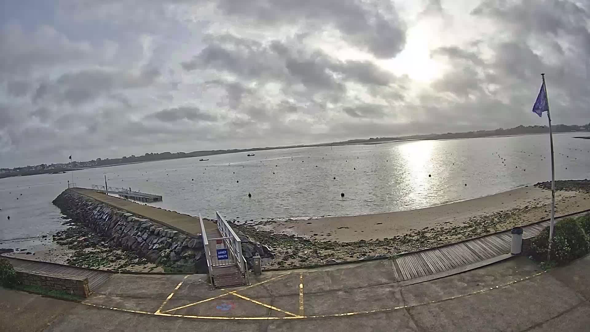 A mostly cloudy day shows a calm body of water with a sandy beach, a stone jetty, and a wooden walkway leading to a small dock.
