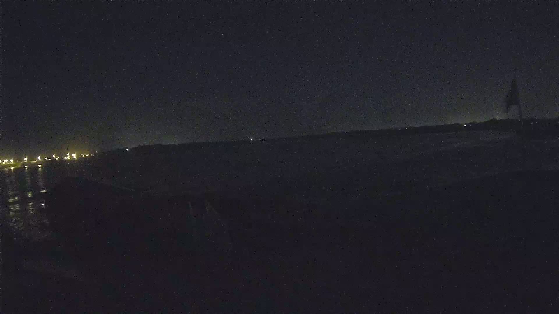 A dark, nighttime scene shows a body of water with a distant shoreline featuring faint lights under a clear, starless sky.