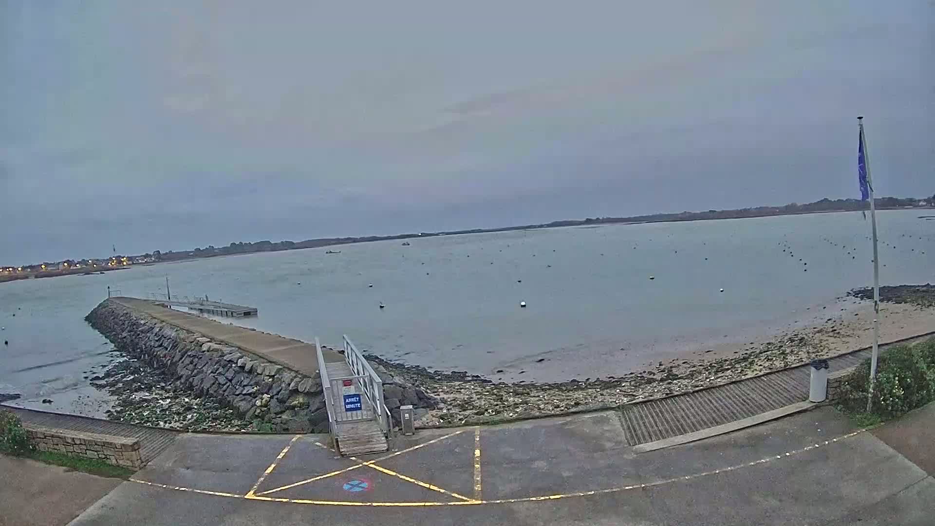 A calm, overcast day reveals a stone jetty leading to a small dock on a body of water, with a sandy shore and distant land visible.