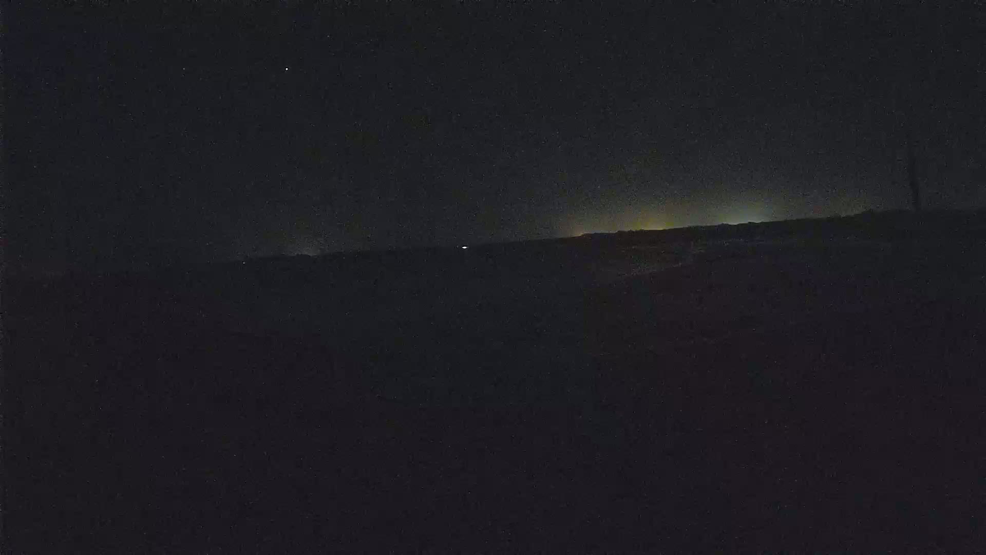 A dark, nighttime landscape shows a faintly lit horizon under a mostly clear, starry sky.
