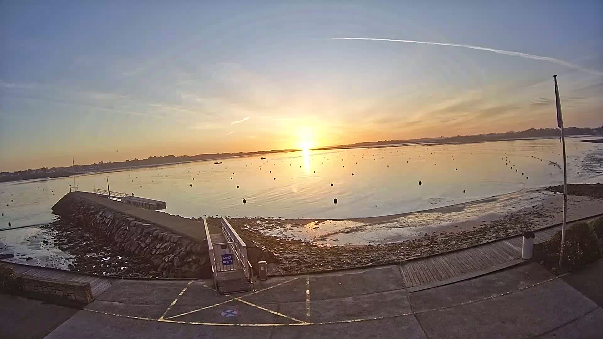 A sunrise over calm water with numerous buoys, a stone jetty, and a paved area leading to a ramp.