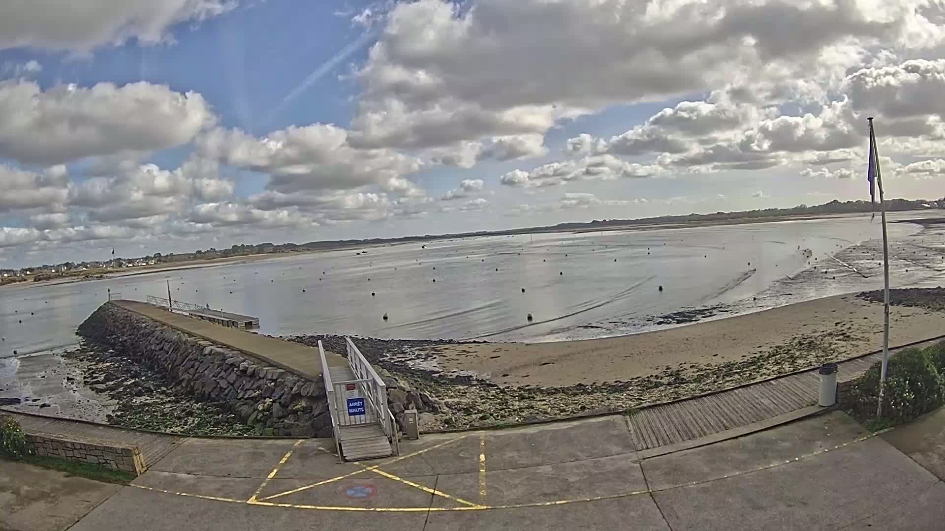 A mostly sunny day with some clouds shows a calm bay with a rocky jetty and a sandy beach.