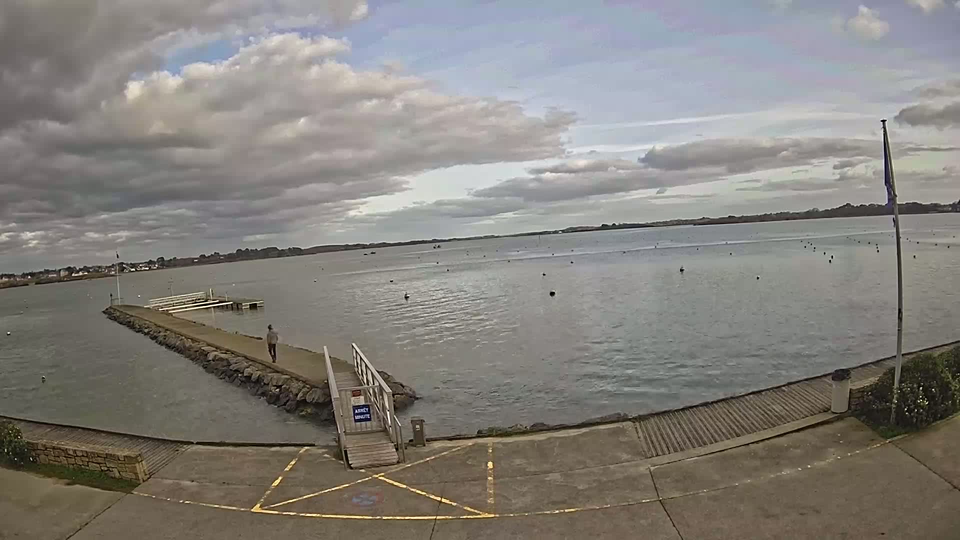 A person walks on a stone jetty extending into a calm body of water under a cloudy sky.