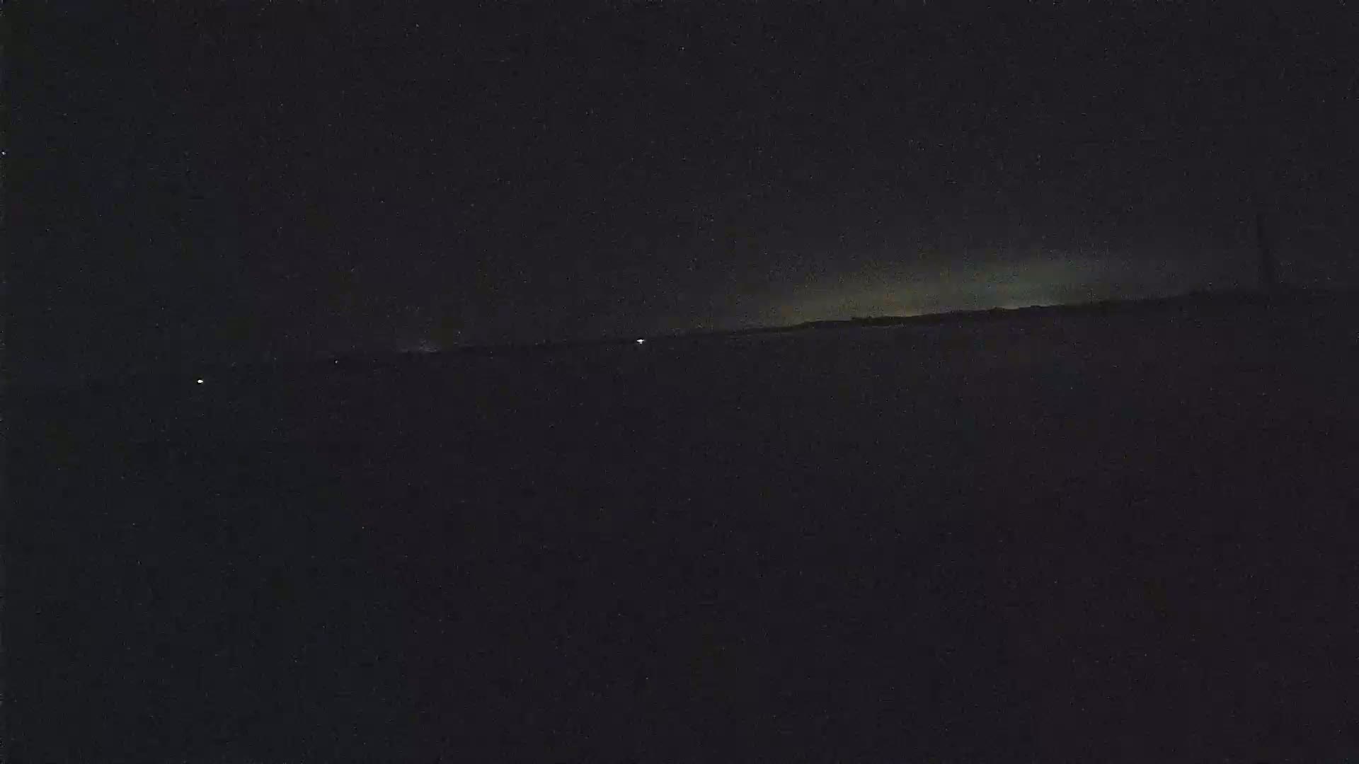 A dark, nighttime landscape shows a dimly lit horizon under a mostly clear sky.