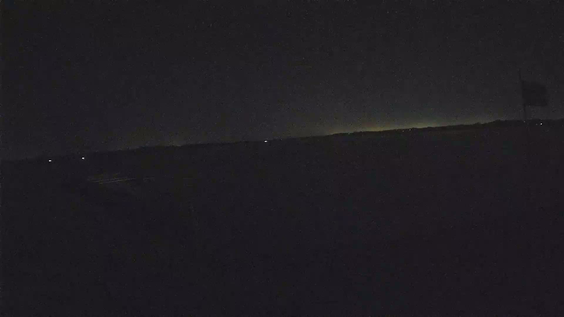 A dark, nighttime landscape shows a faint, light horizon under a mostly clear, star-filled sky.